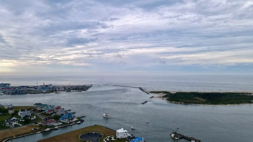 Aerial view over Sinepuxent Bay toward the Ocean City Inlet, with waterfront homes and boats in the foreground